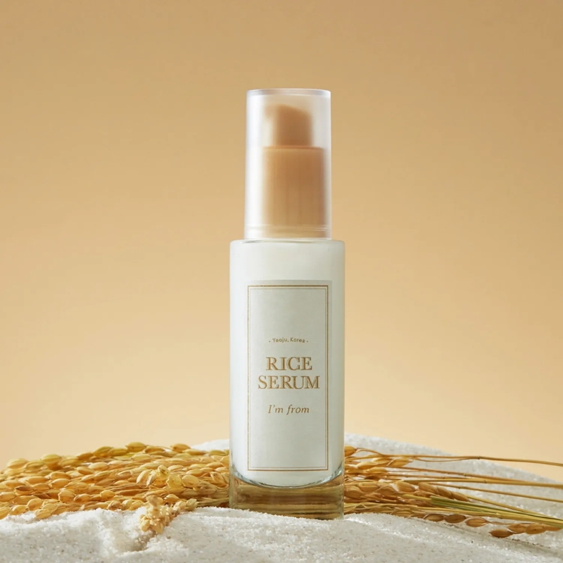 Bottle of rice serum with a pump, standing on white sand with rice grains in foreground.