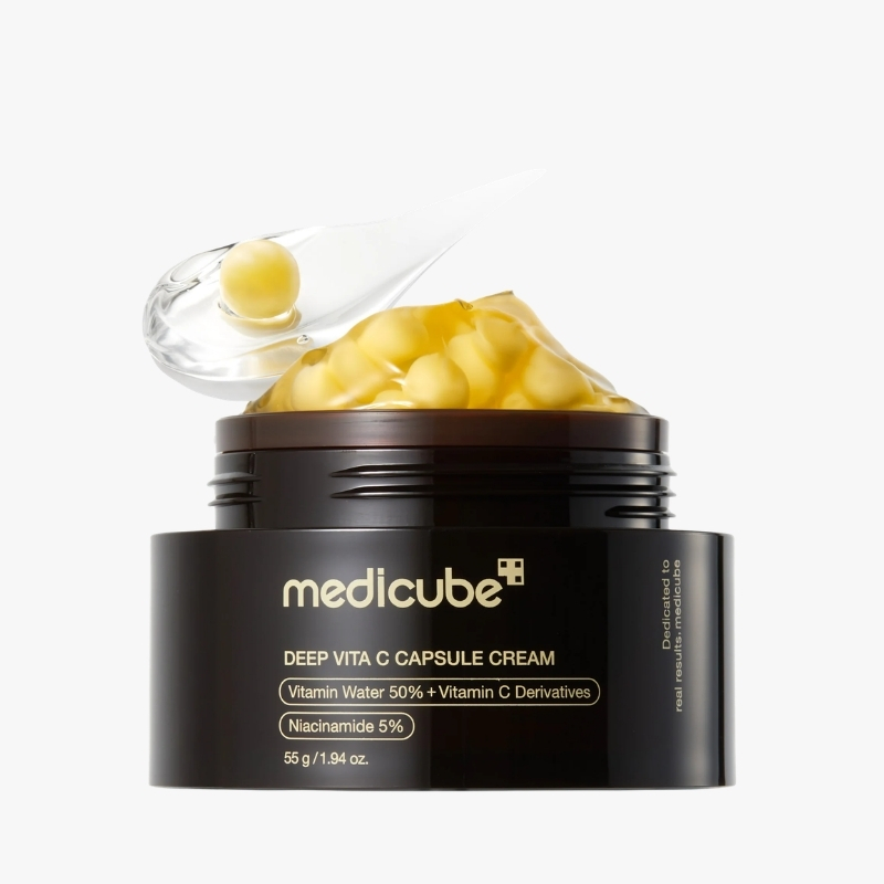 Medicube Deep Vita C Capsule Cream jar with a clear scoop releasing capsules into the open lid, on a white background, black container.