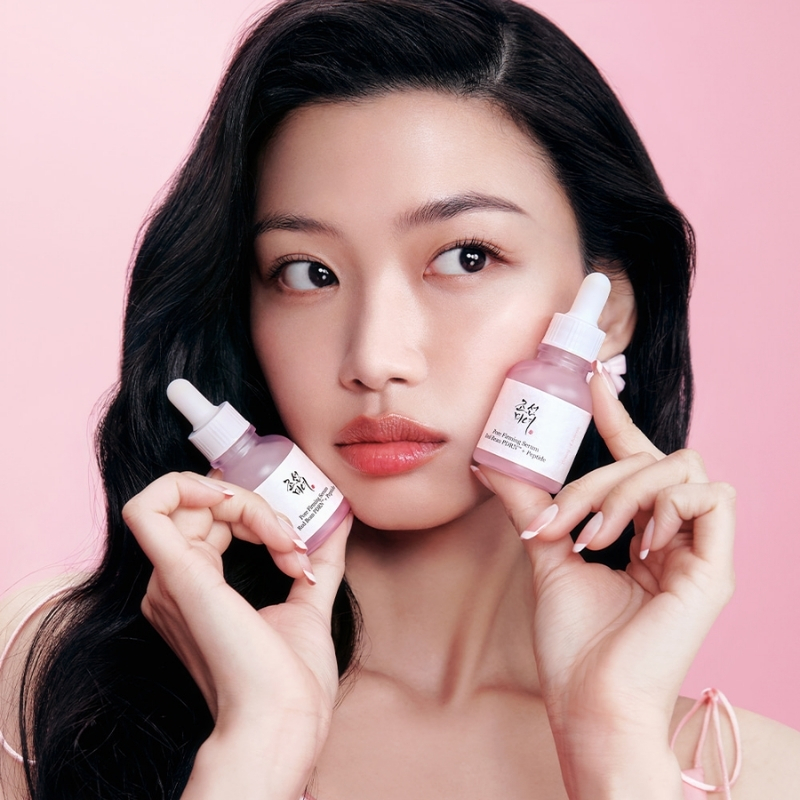 Portrait of a woman with long dark hair holding two pink serum bottles near her face against a pink background for a beauty ad.