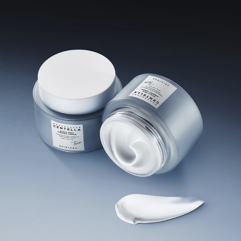 Two frosted glass jars of moisturizer with white lids, one open showing swirled white cream; the label reads 'Centella'.