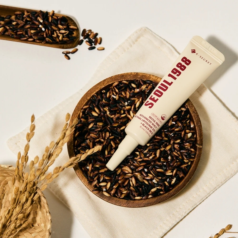 Skincare tube labeled Seoul 1988 resting on a wooden bowl filled with dark brown rice kernels on a beige cloth.