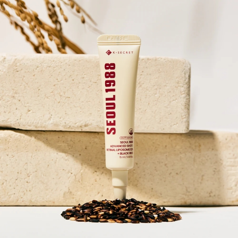 K-Secret Seoul888 cosmetic cream tube upright on a white surface, with a pile of seeds at its base and beige stone blocks in the background.