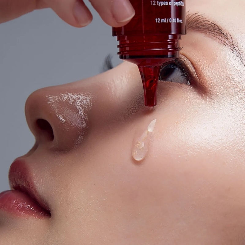 Close-up of a person applying red serum from a dropper near the eye, a droplet forming on the cheek.