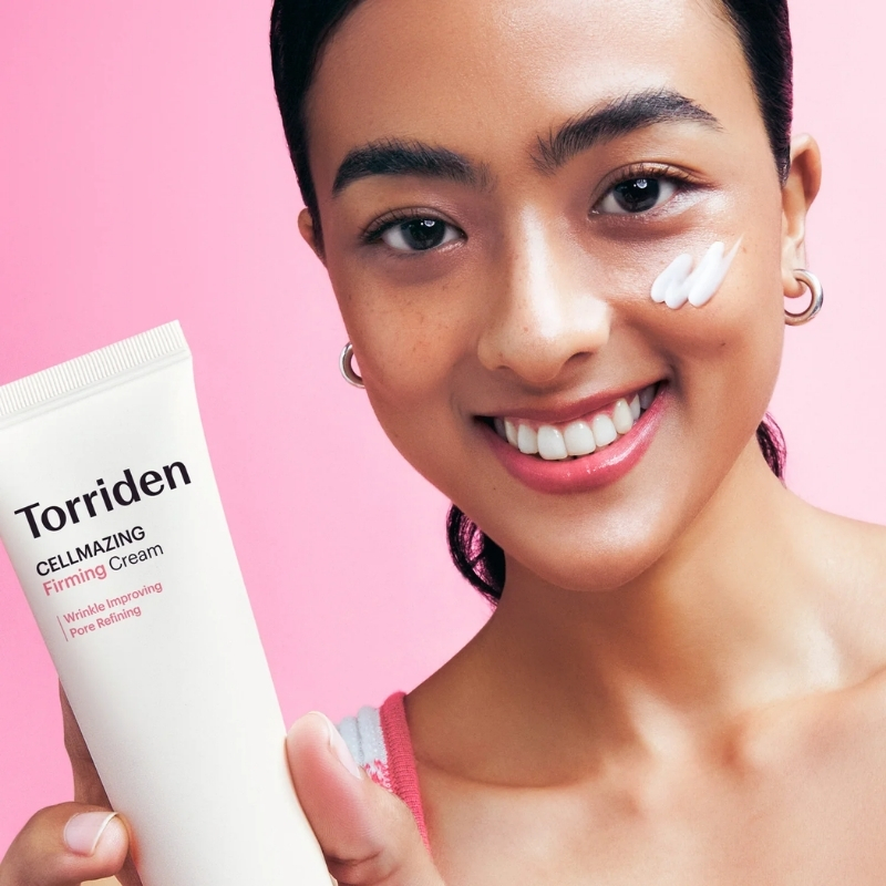 Smiling woman with a dollop of white cream on her cheek holds a Torriden Cellmazing Firming Cream tube against a pink background.