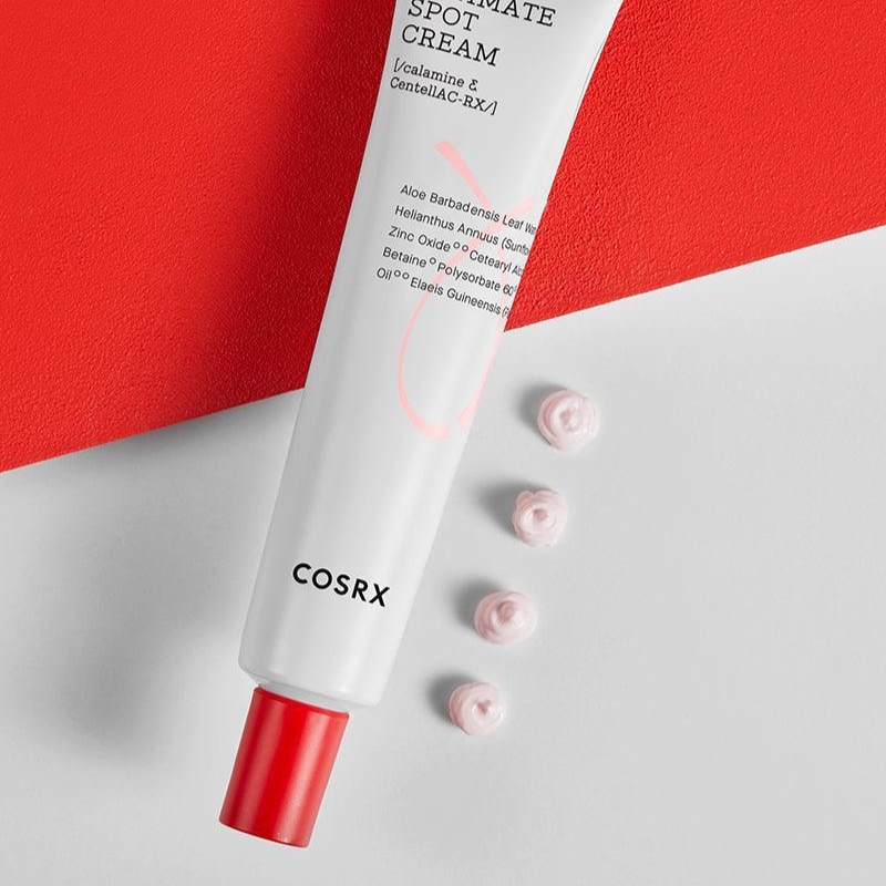 COSRX Ultimate Spot Cream tube with a red cap, angled on a red and white backdrop, with small pink cream swirls beside it.