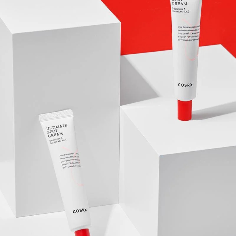 Two white COSRX cosmetic tubes with red caps displayed on white geometric blocks; one reads 'Ultimate Spot Cream' on the label.