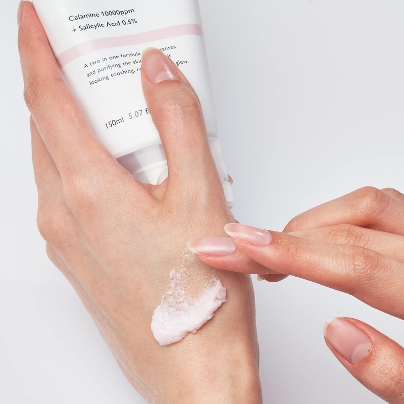 Person applies a dollop of pale pink cream to the back of their forearm from a white skincare tube.